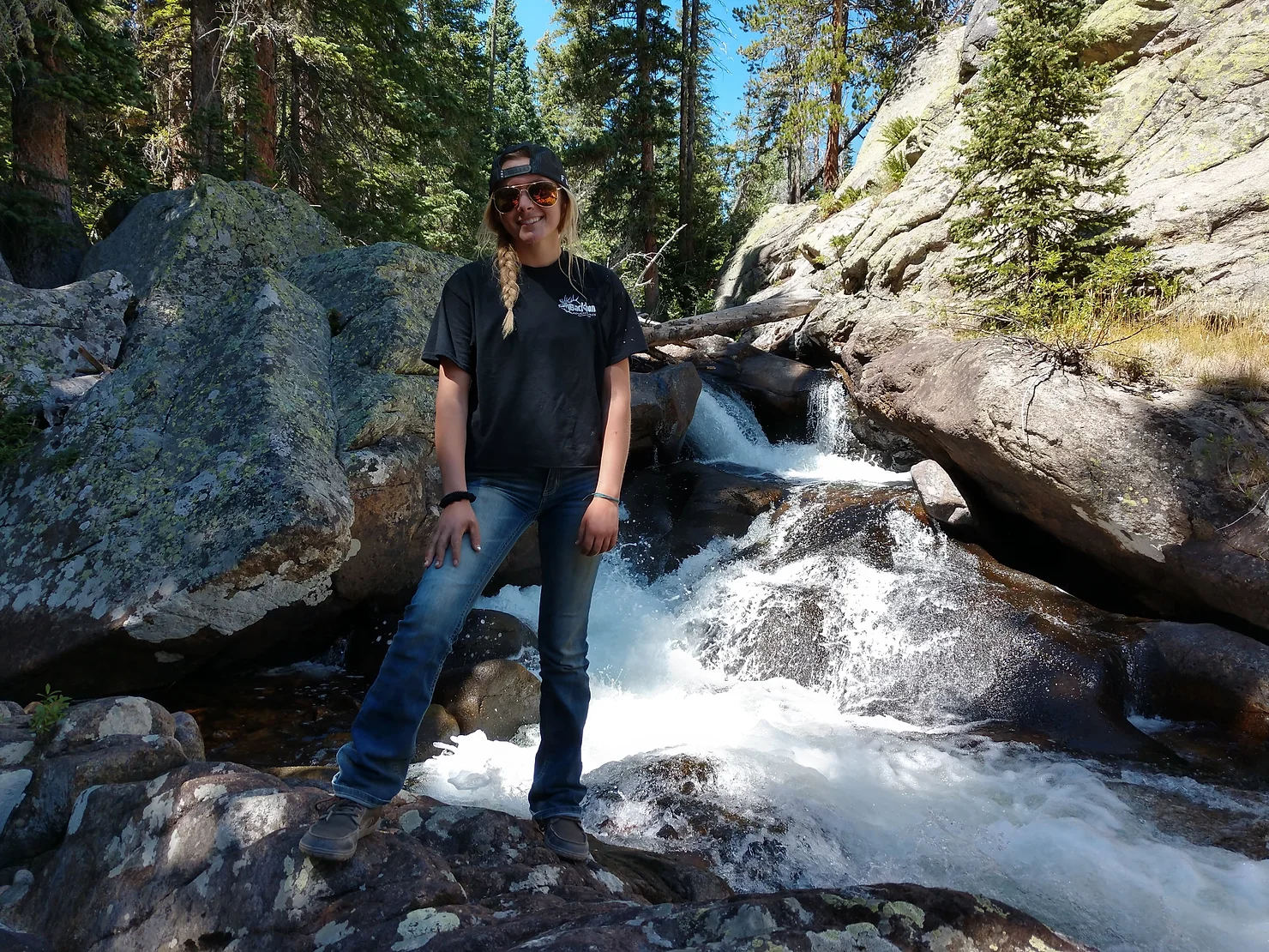 A blond woman with a black ball cap and ponytail stand beside a small water fall in the rocky mountains